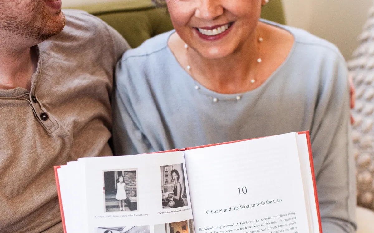 A woman holding open her finished hardcover life story, showing a numbered chapter beside scanned family photographs