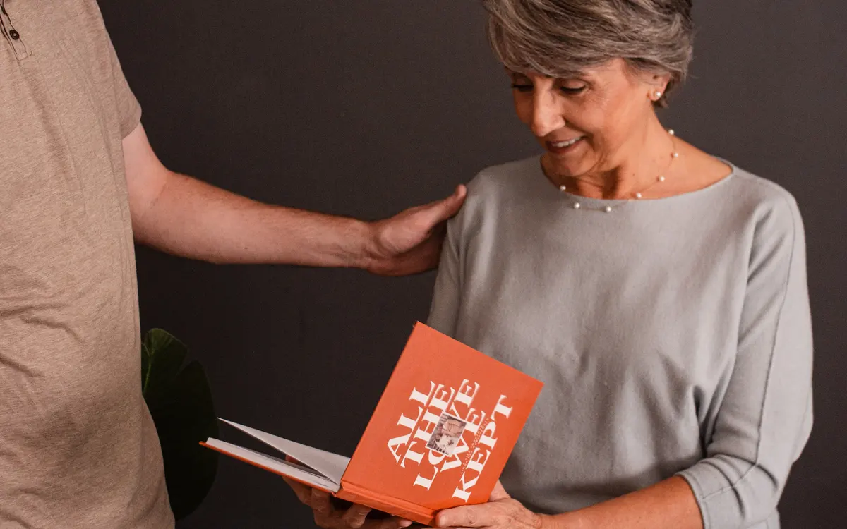 A grown son handing his mother her finished hardcover life story, one hand on her shoulder