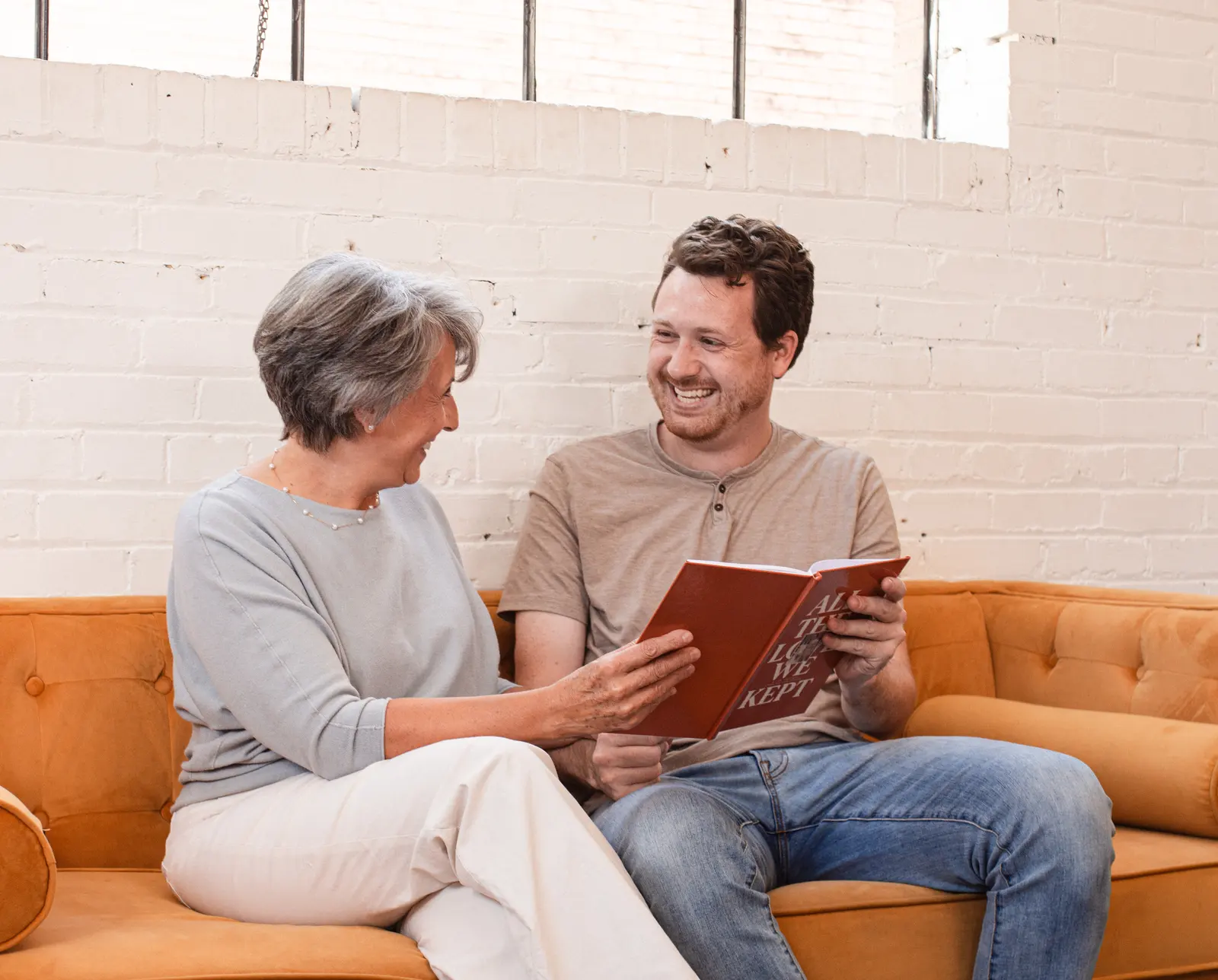 A mother and her grown son laughing together on a sofa, reading her finished hardcover life story