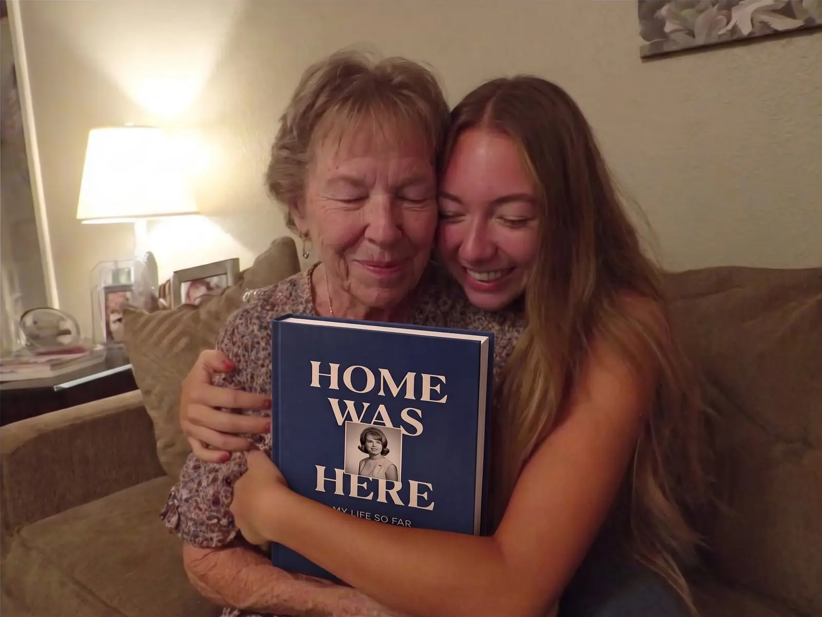 A mother and daughter cheek to cheek on the sofa, hugging her finished Ethos life story hardcover, Home Was Here
